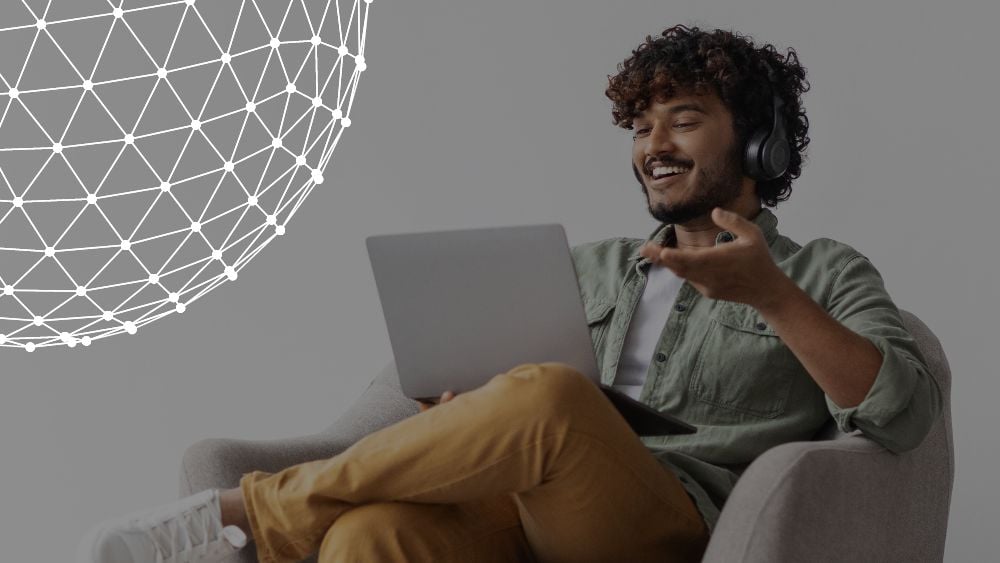 Positive handsome young Indian man sitting in armchair over white studio background, using laptop and headset, having conversation