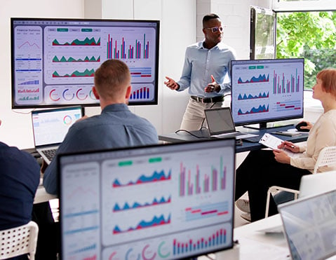 A team of corporate workers sit together and look at statistic KPIs in an office room on screens