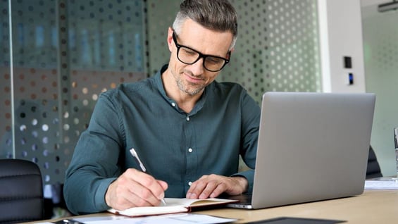 Mature educated man takes notes while being educated remotely via laptop in an home office environment