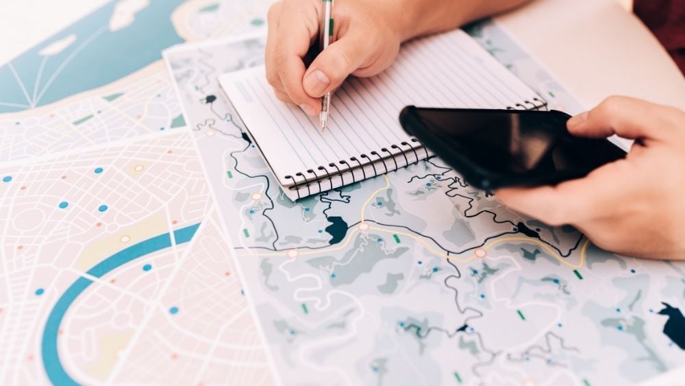 Writing down notes in a notebook with phone in hand and a map on the table
